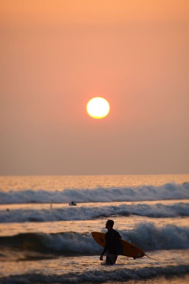A surfer walking into the water at sunset
